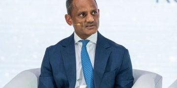 Man in a dark blue suit and light blue tie sits on a stage with a headset microphone, speaking at a panel discussion.