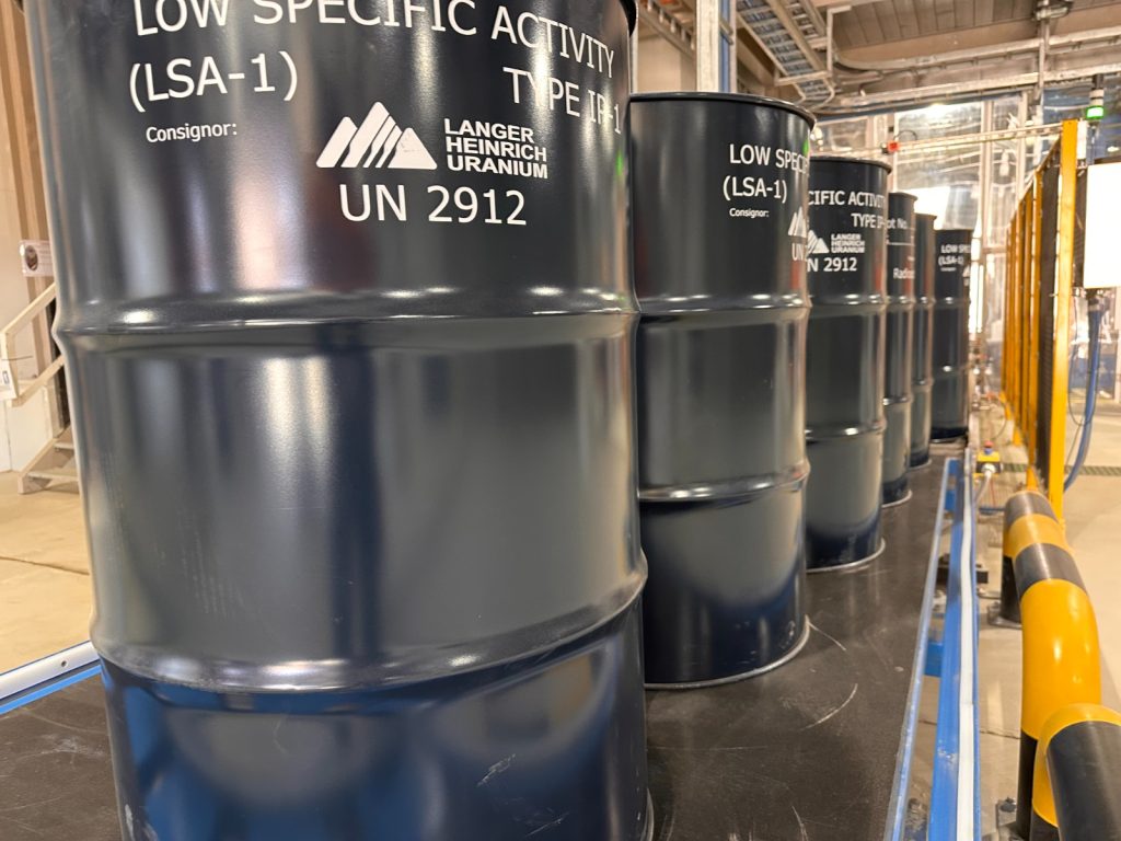 Row of dark blue industrial barrels labeled Low Specific Activity (LSA-1) UN 2912 in a factory setting near safety rails.