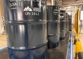 Row of dark blue industrial barrels labeled Low Specific Activity (LSA-1) UN 2912 in a factory setting near safety rails.