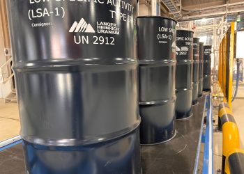 Row of dark blue industrial barrels labeled Low Specific Activity (LSA-1) UN 2912 in a factory setting near safety rails.