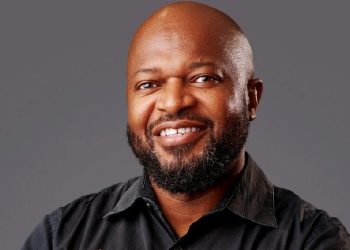 Smiling, bald man with a beard wearing a black shirt against a gray studio backdrop.