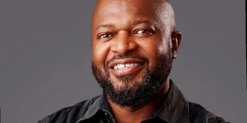 Smiling, bald man with a beard wearing a black shirt against a gray studio backdrop.