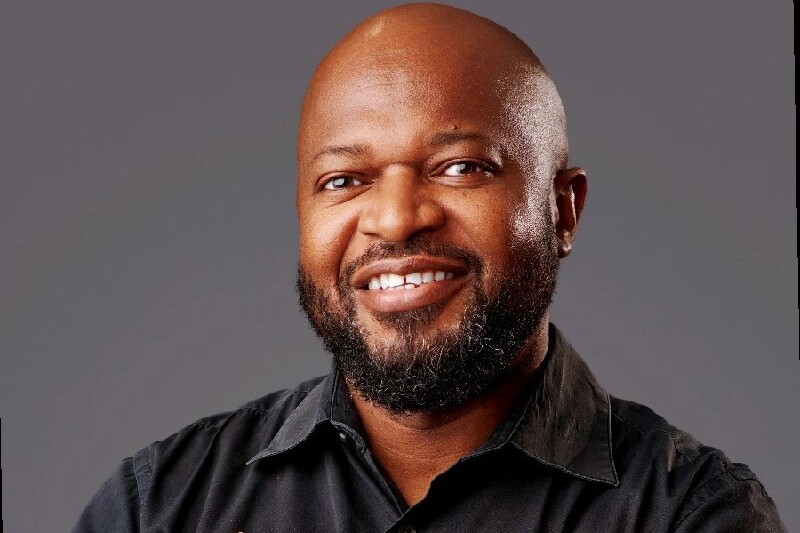 Smiling, bald man with a beard wearing a black shirt against a gray studio backdrop.