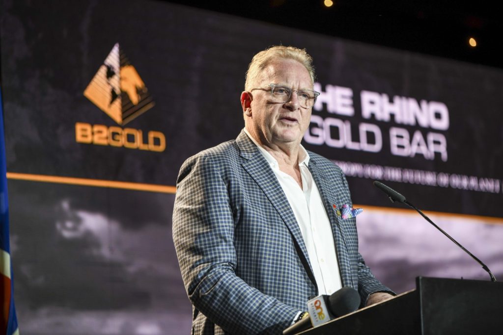 Middle-aged man in a blue checkered blazer speaks at a podium on a stage with a large screen showing a logo in the background.