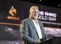 Middle-aged man in a blue checkered blazer speaks at a podium on a stage with a large screen showing a logo in the background.