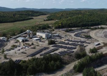 A solar farm with dozens of photovoltaic panels arranged in rows near a small industrial complex amid rocky terrain in a wooded area.