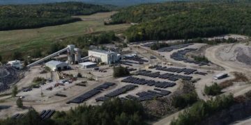 A solar farm with dozens of photovoltaic panels arranged in rows near a small industrial complex amid rocky terrain in a wooded area.