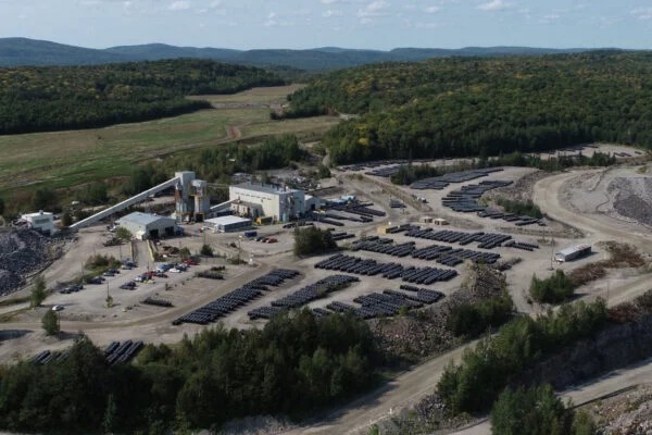 A solar farm with dozens of photovoltaic panels arranged in rows near a small industrial complex amid rocky terrain in a wooded area.