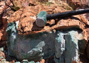 Rock with a greenish turquoise mineral vein and a black hammer resting on top, outdoors among red-brown boulders.