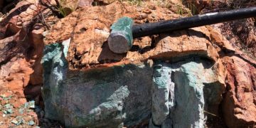Rock with a greenish turquoise mineral vein and a black hammer resting on top, outdoors among red-brown boulders.