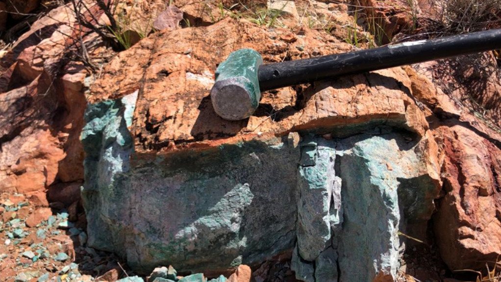 Rock with a greenish turquoise mineral vein and a black hammer resting on top, outdoors among red-brown boulders.