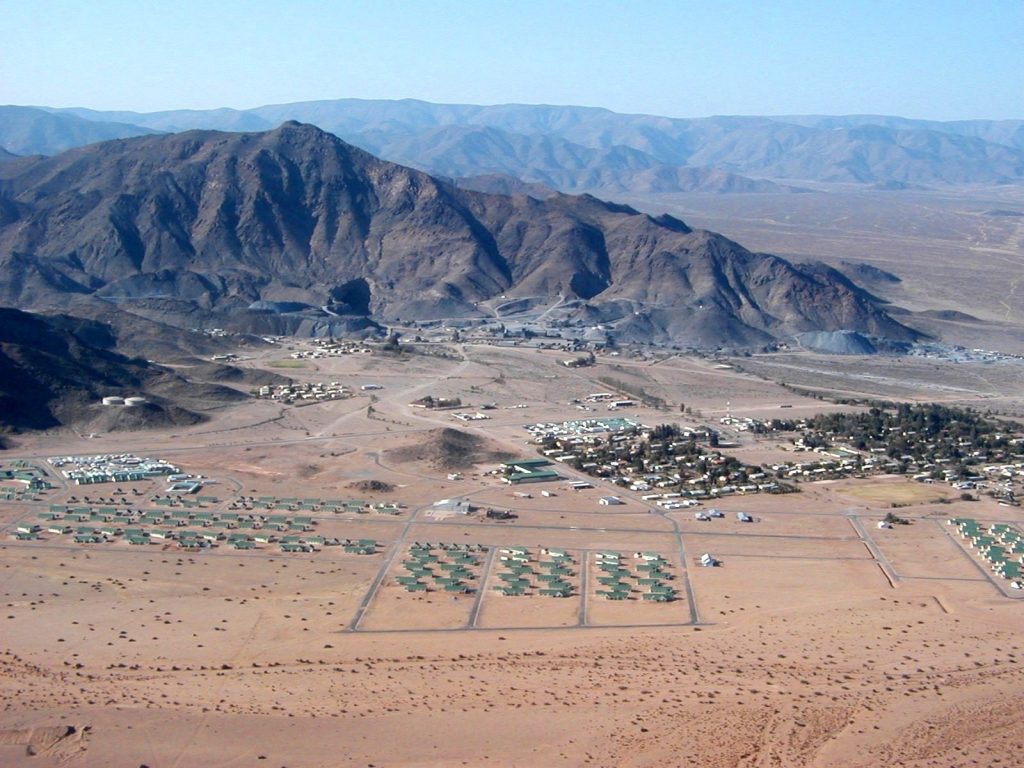 Desert valley with a cluster of green-roofed buildings at the base of rugged mountains, viewed from above.