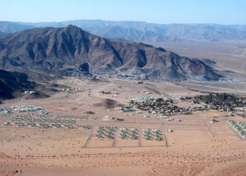 Desert valley with a cluster of green-roofed buildings at the base of rugged mountains, viewed from above.