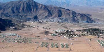 Desert valley with a cluster of green-roofed buildings at the base of rugged mountains, viewed from above.
