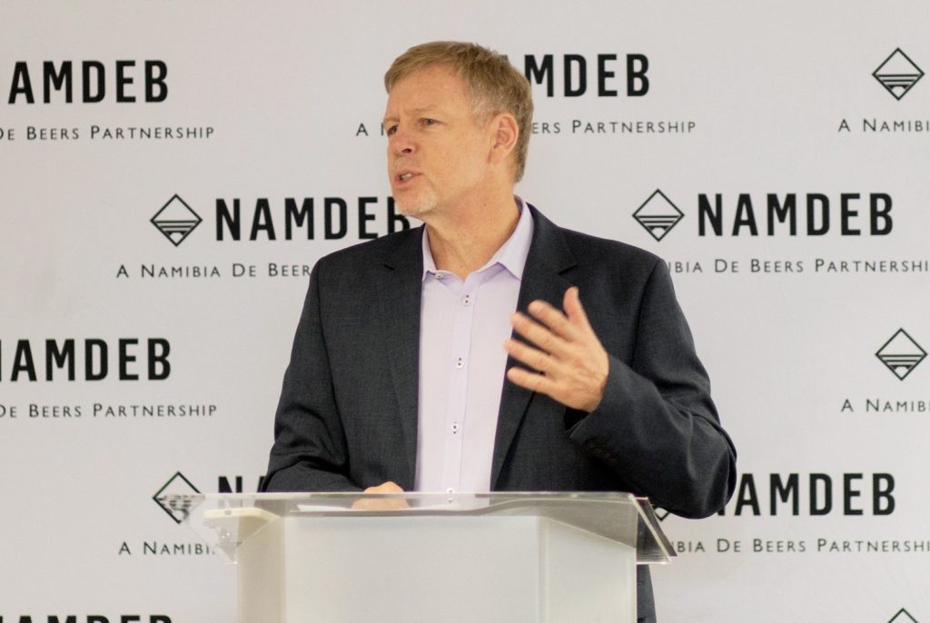 Man in a dark suit speaks at a podium, NAMDEB branding behind him.