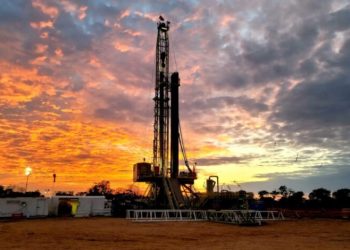 Oil drilling rig on a flat site at sunset, with an orange and purple sky behind it.