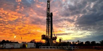 ReconAfrica 2 | Mining and Energy Namibia | Namibia’s Leading Mining & Energy News Oil drilling rig on a flat site at sunset, with an orange and purple sky behind it.