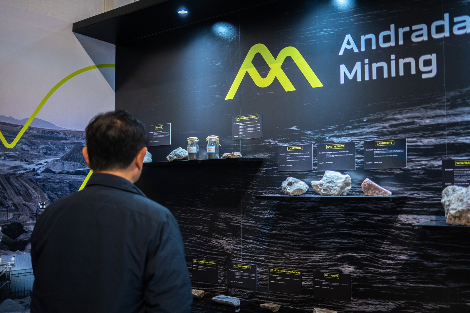 Person in a dark jacket observes a mining exhibit with labeled rock samples on a black wall reading'Andrada Mining'.