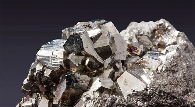 Cluster of metallic cubic pyrite crystals on a rocky matrix with shiny, reflective facets against a dark background.