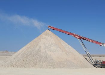 Large mound of light gravel or sand in a desert, with a red conveyor belt depositing material atop the pile.