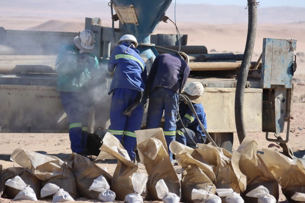 Workers in blue uniforms and white helmets inspect a dusty piece of heavy machinery in a desert worksite, with brown paper bags in the foreground.