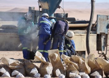 WhatsApp Image 2026-05-19 at 15025 PM | Mining and Energy Namibia | Namibia’s Leading Mining & Energy News Workers in blue uniforms and white helmets inspect a dusty piece of heavy machinery in a desert worksite, with brown paper bags in the foreground.