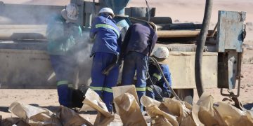 Workers in blue uniforms and white helmets inspect a dusty piece of heavy machinery in a desert worksite, with brown paper bags in the foreground.