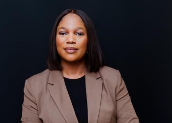 Professional woman in a tan blazer and black top, looking confidently at the camera against a dark background.