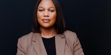 Professional woman in a tan blazer and black top, looking confidently at the camera against a dark background.
