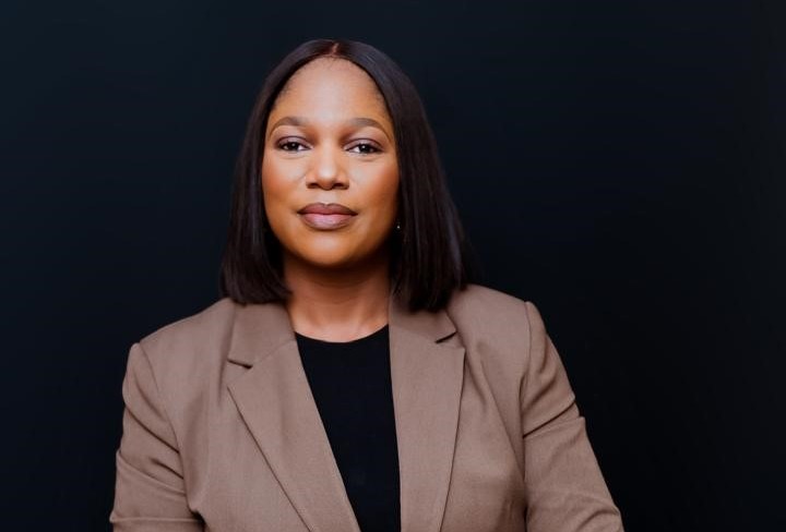 Professional woman in a tan blazer and black top, looking confidently at the camera against a dark background.