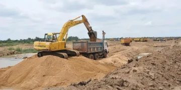 Yellow excavator loading sand into a dump truck at a dirt construction site, with more equipment lined up in the distance