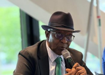 A man in a dark suit, teal tie, and fedora speaks into a handheld microphone at a conference table, with a glass of water nearby.