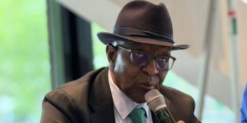 A man in a dark suit, teal tie, and fedora speaks into a handheld microphone at a conference table, with a glass of water nearby.