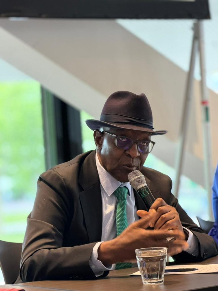 kaire Mbuende | Mining and Energy Namibia | Namibia’s Leading Mining & Energy News A man in a dark suit, teal tie, and fedora speaks into a handheld microphone at a conference table, with a glass of water nearby.