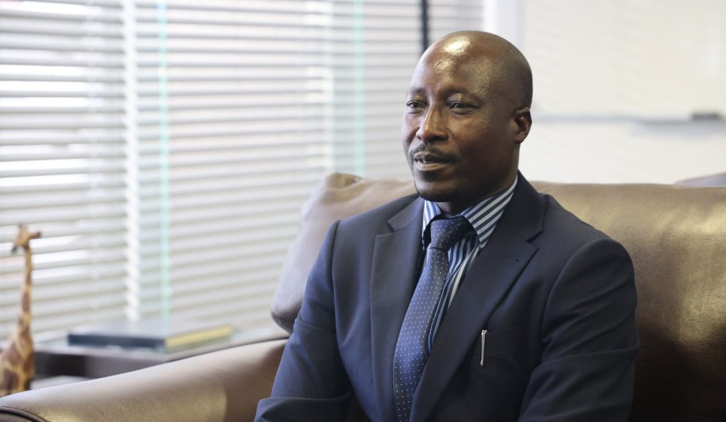 Sakarias Uunona | Mining and Energy Namibia | Namibia’s Leading Mining & Energy News Professional man in a navy suit and striped shirt seated on a leather couch in an office setting