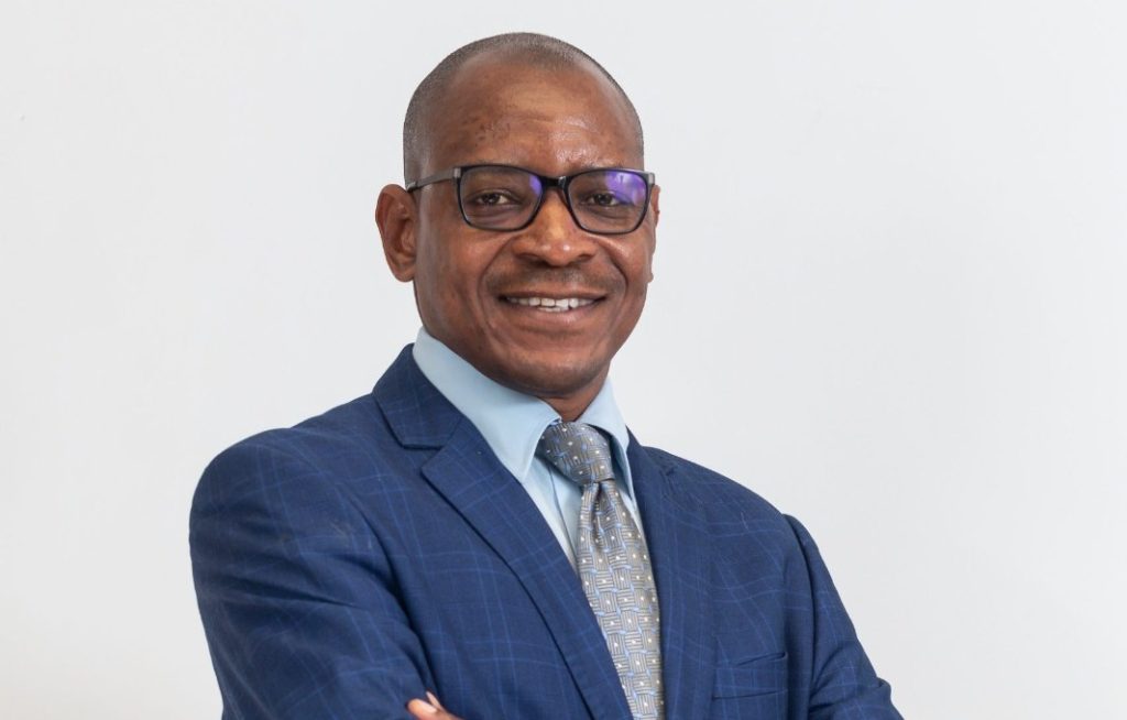 GEORGE BOTSHIWE3 | Mining and Energy Namibia | Namibia’s Leading Mining & Energy News Smiling man in a blue suit, light blue shirt, and patterned gray tie, wearing glasses, against a white background.
