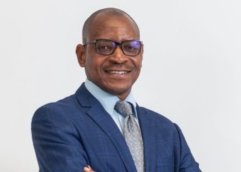 Smiling man in a blue suit, light blue shirt, and patterned gray tie, wearing glasses, against a white background.