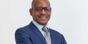 Smiling man in a blue suit, light blue shirt, and patterned gray tie, wearing glasses, against a white background.