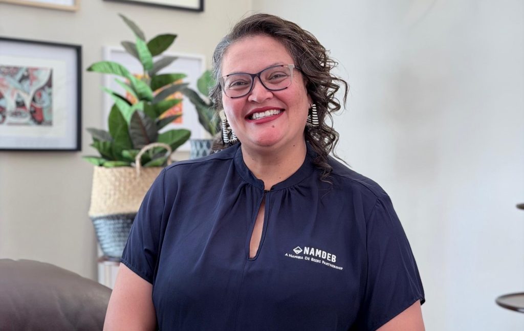 Smiling woman with glasses in a navy blouse, standing indoors with a plant in the background.