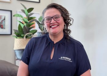 Smiling woman with glasses in a navy blouse, standing indoors with a plant in the background.