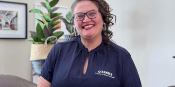 Smiling woman with glasses in a navy blouse, standing indoors with a plant in the background.