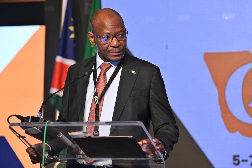 George Botshiwe | Mining and Energy Namibia | Namibia’s Leading Mining & Energy News Man in a dark suit and red tie speaks at a podium with microphones, wearing a lanyard and a flag pin on his lapel.