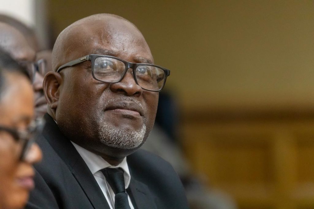 Close-up of an older Black man wearing glasses and a dark suit at a formal event.