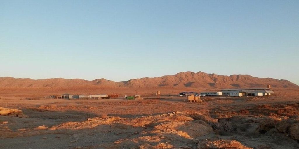 FORSYS | Mining and Energy Namibia | Namibia’s Leading Mining & Energy News Desert settlement with low buildings and storage containers on a rocky plain, mountains in the distance at sunset.