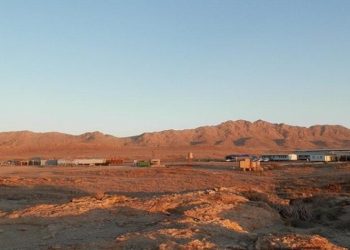 Desert settlement with low buildings and storage containers on a rocky plain, mountains in the distance at sunset.