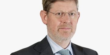 Portrait of a middle-aged man with glasses, gray beard, wearing a dark suit and light blue shirt with a teal tie, looking at the camera.