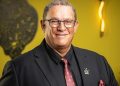 Romeo Nel | Mining and Energy Namibia | Namibia’s Leading Mining & Energy News Smiling man in a dark suit and pink tie, wearing glasses and a lapel pin, posed against a yellow background.