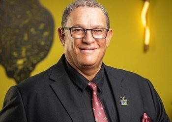 Smiling man in a dark suit and pink tie, wearing glasses and a lapel pin, posed against a yellow background.