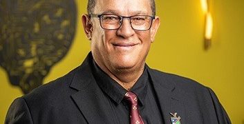 Smiling man in a dark suit and pink tie, wearing glasses and a lapel pin, posed against a yellow background.