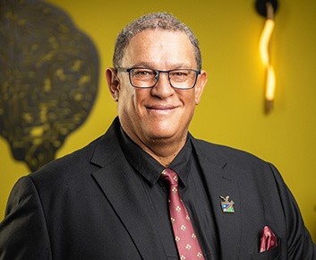 Smiling man in a dark suit and pink tie, wearing glasses and a lapel pin, posed against a yellow background.